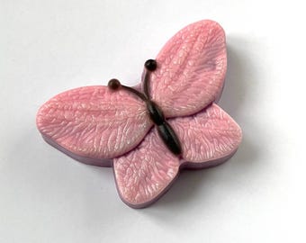 May include: A pink butterfly-shaped decorative item with detailed wing textures. The butterfly has a dark brown body and antennae. The wings are a soft pink with subtle shading, and the item appears to be made of a solid material.