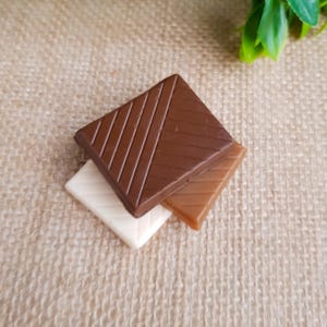 May include: Three square chocolates in different shades of brown and white are stacked on top of each other. The top chocolate is dark brown with diagonal lines. The middle chocolate is light brown, and the bottom one is white.