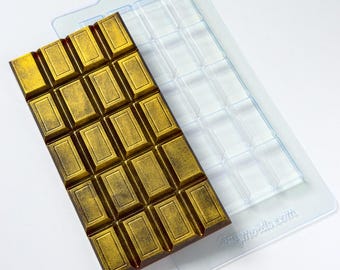 May include: A gold-colored chocolate bar with a rectangular shape, resting next to a clear plastic mold. The chocolate bar has a textured surface and is divided into rectangular sections. The mold is designed to create the same shape.