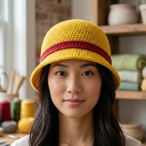 May include: A yellow crochet bucket hat with a red band. The hat has a rounded crown and a slightly downward-sloping brim. The hat is worn by a person with dark hair.