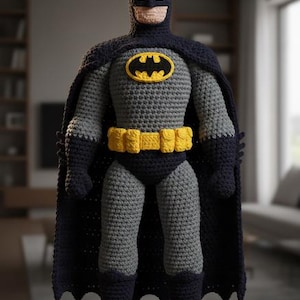 Puede incluir: Muñeco de Batman hecho a mano con ganchillo. La figura es principalmente gris con una capa, capucha y botas negras. El icónico símbolo del murciélago amarillo está en el pecho y un cinturón utilitario amarillo en la cintura.