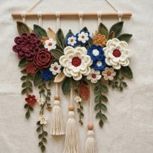 May include: A wall hanging featuring crocheted flowers and leaves in shades of burgundy, cream, blue, and green. The arrangement is suspended from a horizontal wooden dowel by cream-colored cords, with three hanging tassels and wooden beads.