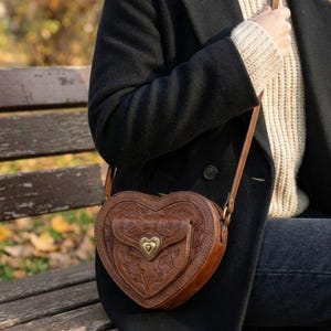 Puede incluir: Bolso bandolera de cuero marrón en forma de corazón con detalles florales en relieve. El bolso tiene un pequeño bolsillo frontal con un cierre en forma de corazón y una correa larga y ajustable.