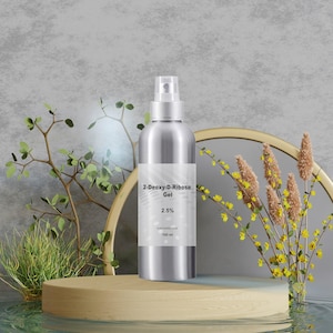 May include: A silver spray bottle with a white label displaying "2-Deoxy-D-Ribose Gel 2.5%" and "100 ml". The bottle is on a light brown wooden platform with greenery and a gold-framed mirror in the background. The background is a light gray.