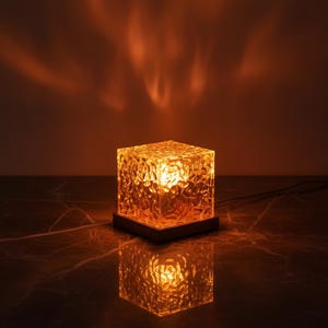 Puede incluir: Lámpara cúbica texturizada que emite un cálido brillo ámbar. La lámpara se asienta sobre una base de madera oscura, creando una luz suave y un reflejo. La superficie del cubo presenta un patrón irregular, lo que añade a su diseño único.