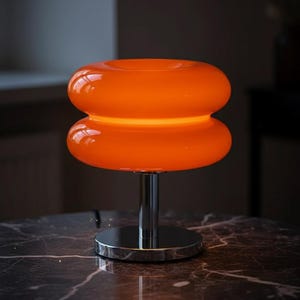 Peut inclure: Lampe de table orange vif avec un abat-jour arrondi empilé. La lampe a une base et une tige en chrome poli, et elle repose sur une surface en marbre foncé. L'abat-jour de la lampe émet une lueur interne.