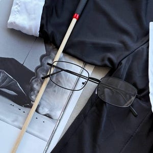 May include: Black rectangular eyeglasses with thin black frames rest on a black garment. A wooden stick with a black handle and red accent is also present. A white collared shirt is visible in the background.