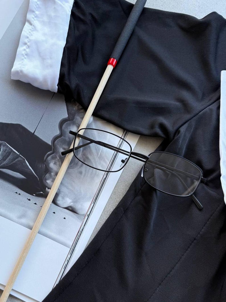 May include: Black rectangular eyeglasses with thin black frames rest on a black garment. A wooden stick with a black handle and red accent is also present. A white collared shirt is visible in the background.