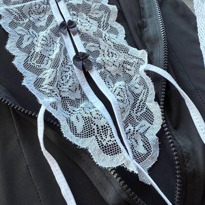 May include: Black jacket with a white lace collar and small black bows. The collar is detailed with intricate lace patterns and white ribbons. A zipper runs down the front of the jacket.