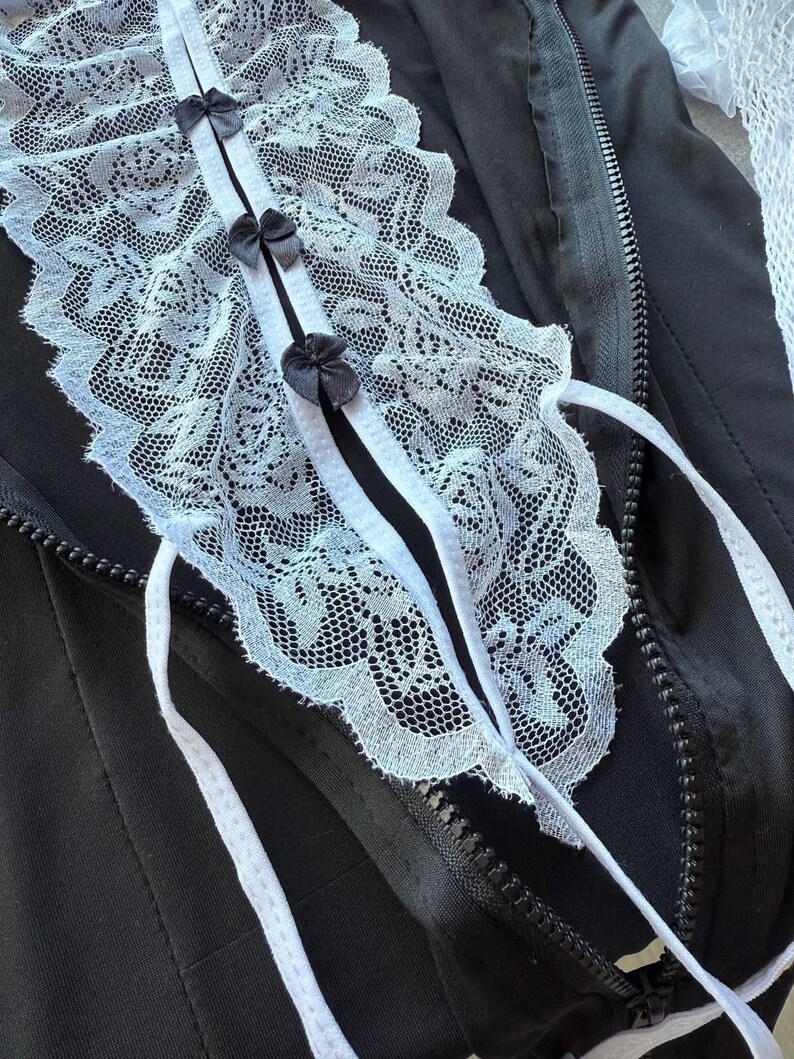 May include: Black jacket with a white lace collar and small black bows. The collar is detailed with intricate lace patterns and white ribbons. A zipper runs down the front of the jacket.