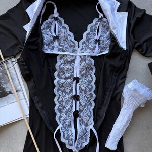 May include: Black lingerie set with white lace detailing. The set includes a top, a bodice, and a pair of white lace gloves. The bodice has black bows and a zipper closure. The top has white lace trim and black bows.