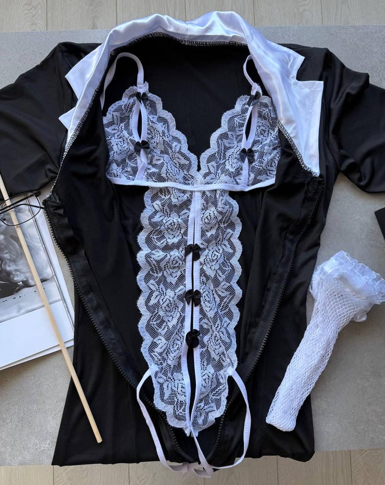 May include: Black lingerie set with white lace detailing. The set includes a top, a bodice, and a pair of white lace gloves. The bodice has black bows and a zipper closure. The top has white lace trim and black bows.