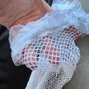 May include: Close-up of a white fishnet stocking with a ruffled, sheer trim. The stocking is held up, revealing the open weave of the fabric. The background is a dark gray shirt.