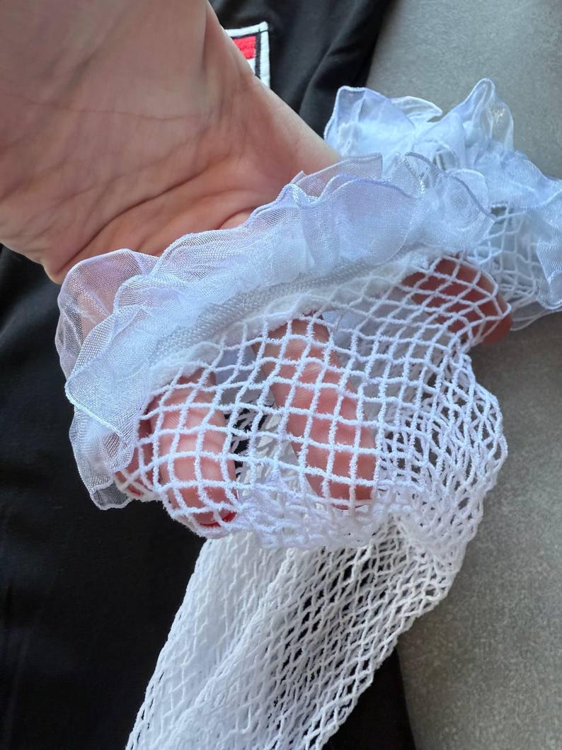 May include: Close-up of a white fishnet stocking with a ruffled, sheer trim. The stocking is held up, revealing the open weave of the fabric. The background is a dark gray shirt.