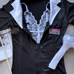 May include: Black lingerie set with a white lace bodice and a black jacket with white lapels. The jacket has a zipper closure and a "SCHOOL TEACHER" patch. Includes a pair of glasses and a white fishnet sleeve.