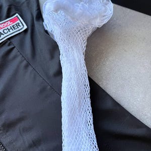 May include: A white, net-like accessory with a ruffled top, possibly a hair accessory or part of a costume, is displayed against a black jacket. The item is made of a delicate, open-weave material.