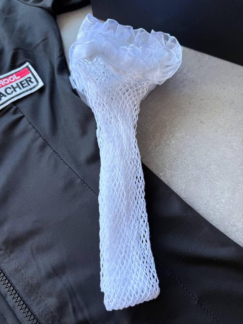 May include: A white, net-like accessory with a ruffled top, possibly a hair accessory or part of a costume, is displayed against a black jacket. The item is made of a delicate, open-weave material.