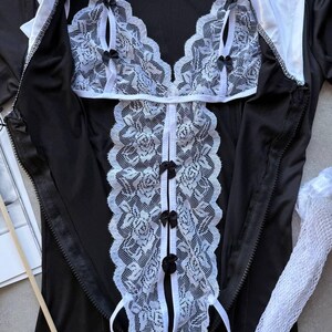 May include: Black lingerie set with white lace detailing. The set includes a top with a halter neck and a matching bottom. The lace features a floral pattern and is accented with small black bows. The outfit has a zipper closure and white collar.