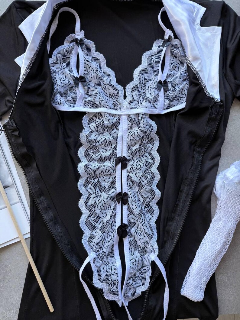 May include: Black lingerie set with white lace detailing. The set includes a top with a halter neck and a matching bottom. The lace features a floral pattern and is accented with small black bows. The outfit has a zipper closure and white collar.