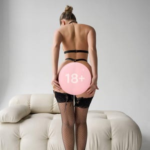 Może przedstawiać: Osoba w czarnej bieliźnie, pończochach z siatki i butach na wysokim obcasie, trzymająca r&oacute;żowy balon z napisem "18+". Tło obejmuje białą sofę i gładką białą ścianę.