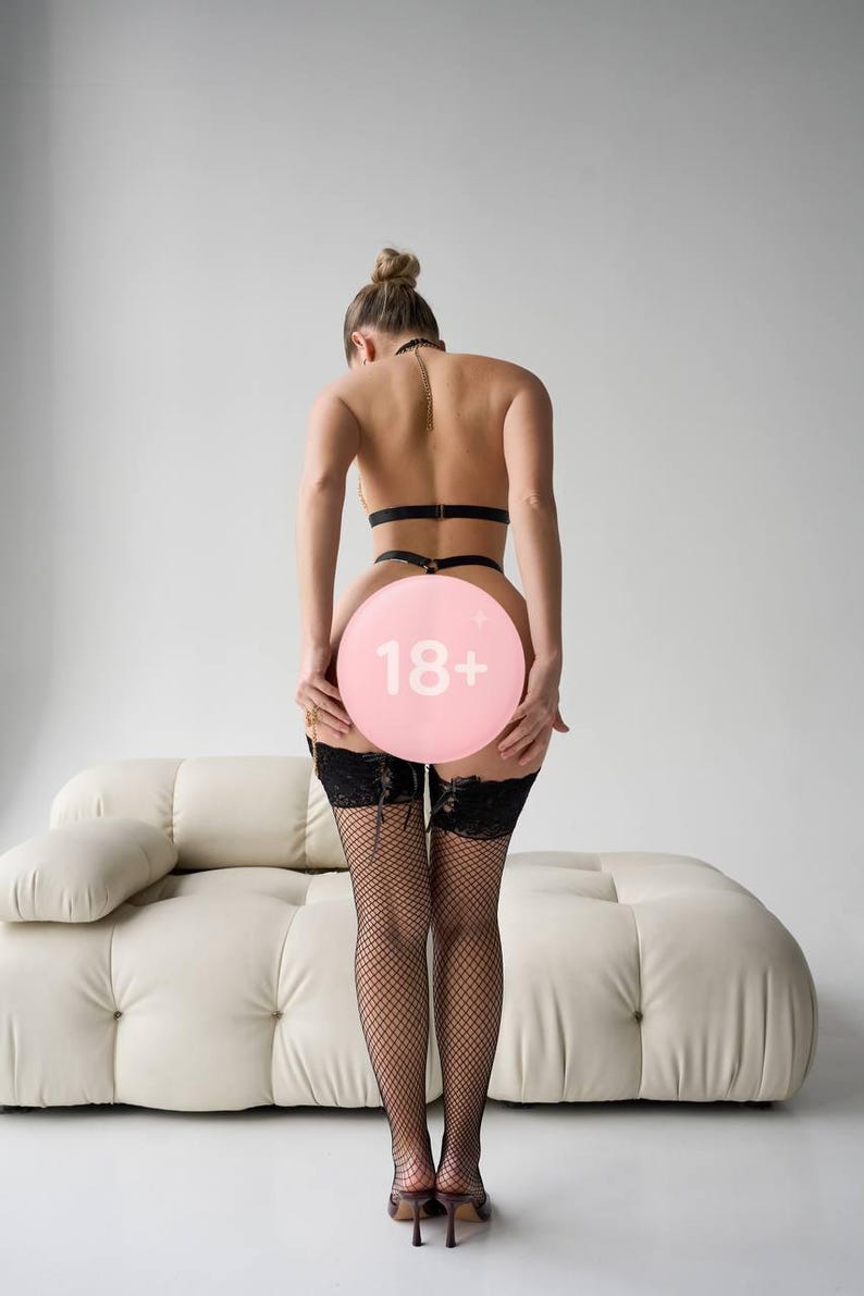 Może przedstawiać: Osoba w czarnej bieliźnie, pończochach z siatki i butach na wysokim obcasie, trzymająca r&oacute;żowy balon z napisem "18+". Tło obejmuje białą sofę i gładką białą ścianę.
