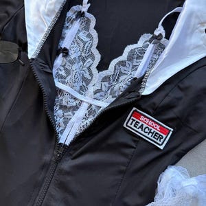 May include: Black school teacher costume with a white lace bralette and collar. The costume has a zipper down the front and a patch that says "SCHOOL TEACHER".