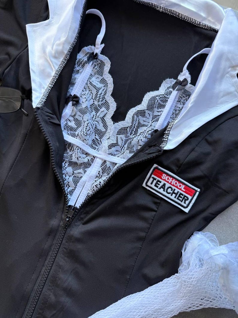 May include: Black school teacher costume with a white lace bralette and collar. The costume has a zipper down the front and a patch that says "SCHOOL TEACHER".