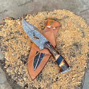 Puede incluir: Un cuchillo de acero de Damasco con una hoja estampada y un mango con tonos marrones y negros. El cuchillo se muestra con una funda de cuero marrón sobre una superficie de madera.