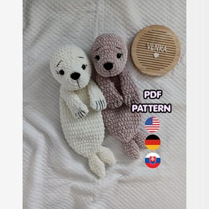 May include: Two handmade crochet seal plushies, one white and one mauve, with black eyes and noses. A wooden coaster with the name "VENKA" is next to the seals. The image also includes the text "PDF PATTERN" and flags.