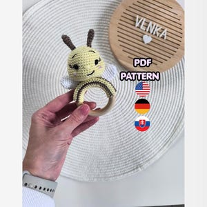 May include: A crocheted bee-shaped baby rattle with a wooden ring. The bee is yellow with brown stripes and antennae, and white wings. The text "PDF PATTERN" is visible, along with the name "VENKA" on a wooden board.