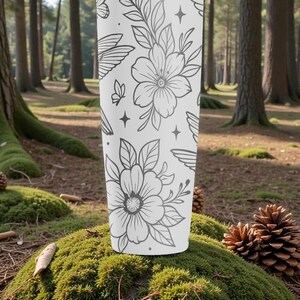 Pu&ograve; includere: Un bicchiere bianco con un motivo floreale e a farfalle grigio. Il design include fiori dettagliati, foglie, farfalle, ali e stelle. Il bicchiere cilindrico &egrave; posto su muschio in un ambiente forestale.