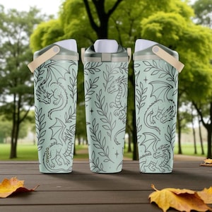 May include: Three water bottles with a light green background and a dragon and leaf design. The bottles have a white top, a tan handle, and the brand name "owala" printed on the side. The bottles are on a wooden table with fall leaves.