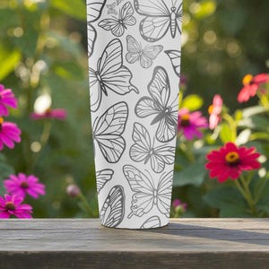 May include: A white tumbler with a repeating pattern of gray butterfly illustrations. The tumbler is cylindrical and stands on a wooden surface, with a blurred background of pink flowers and green foliage.