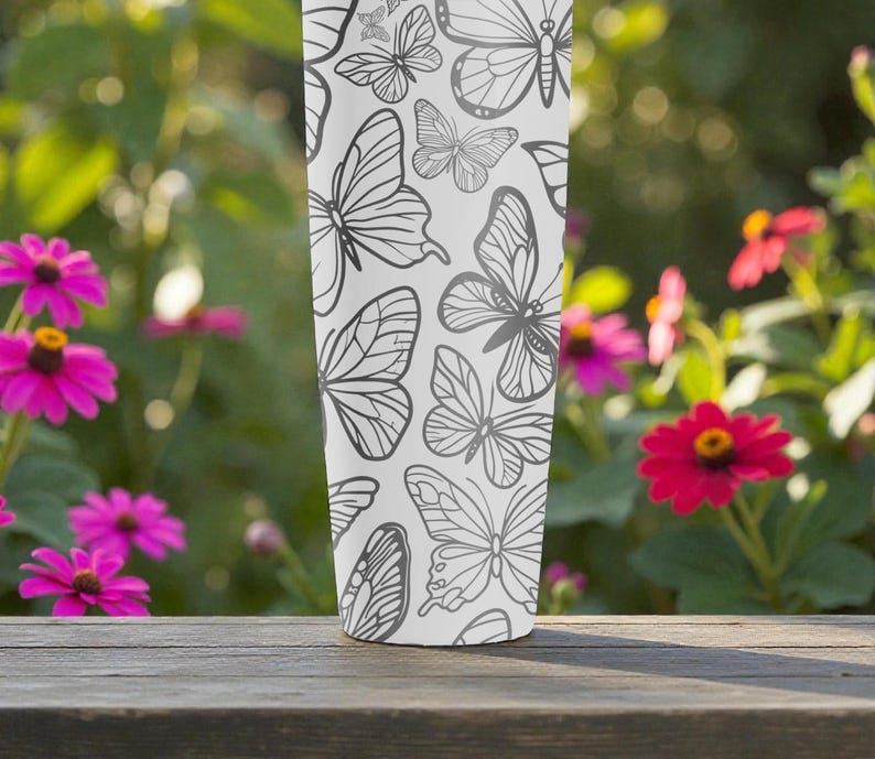 May include: A white tumbler with a repeating pattern of gray butterfly illustrations. The tumbler is cylindrical and stands on a wooden surface, with a blurred background of pink flowers and green foliage.