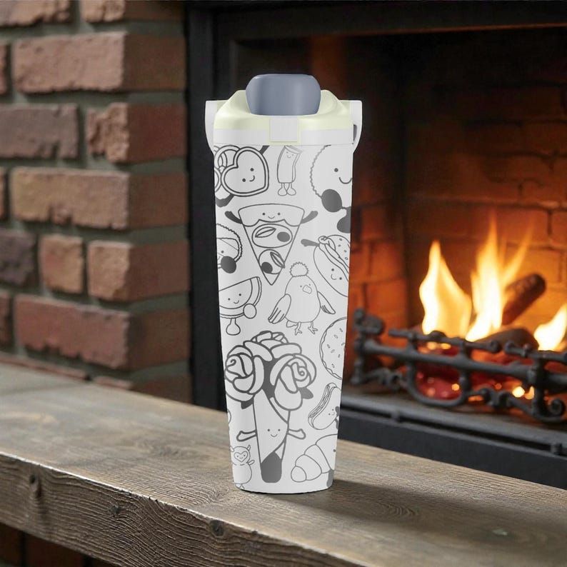 May include: A white travel mug with a gray lid and a whimsical black and white food-themed pattern. The design includes illustrations of pizza slices, roses, and other food items. The mug is set against a backdrop of a brick fireplace.