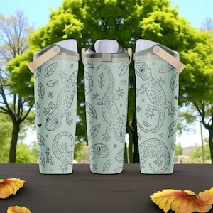 Op de afbeelding: Drie lichtgroene waterflessen met een herhalend zwart lijnpatroon van gekko's, bladeren en kleine hartjes. Elke fles heeft een witte en beige dop met een draaglus. De merknaam Owala staat verticaal op de zijkant van de flessen gedrukt.