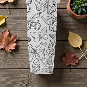 May include: A white tumbler with a black butterfly pattern. The tumbler is surrounded by autumn leaves and a small potted succulent on a wooden surface. The butterflies are in various sizes and designs.