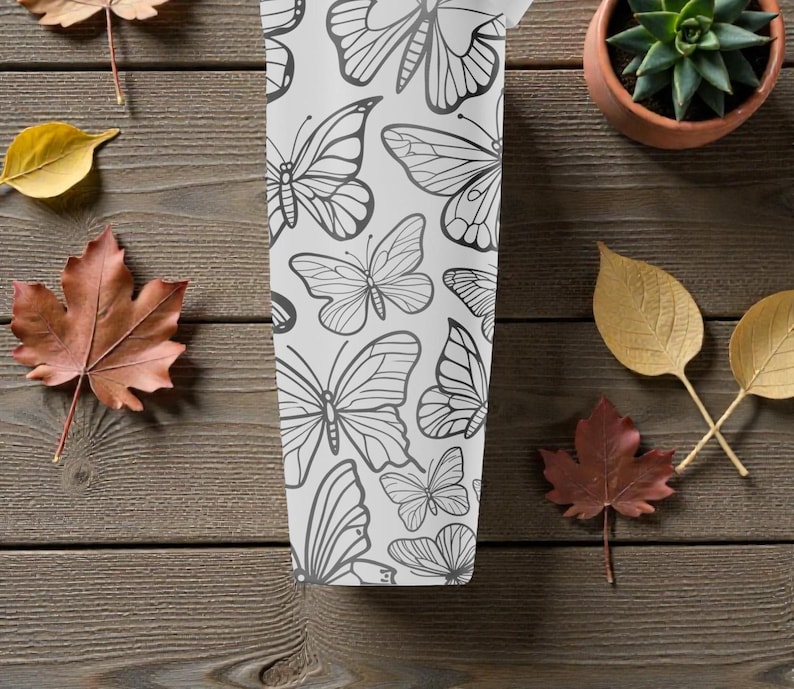 May include: A white tumbler with a black butterfly pattern. The tumbler is surrounded by autumn leaves and a small potted succulent on a wooden surface. The butterflies are in various sizes and designs.