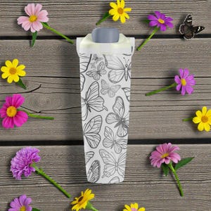 May include: A white tumbler with a gray lid, adorned with a black butterfly pattern. The tumbler is surrounded by colorful flowers on a wooden surface, creating a nature-inspired theme.