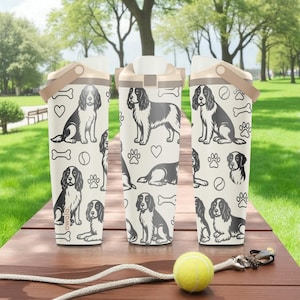 Puede incluir: Tres botellas de agua blancas con un estampado de perros con spaniels en blanco y negro, huellas de patas y huesos. Cada botella tiene un asa y tapa beiges. Una pelota de tenis y una correa descansan sobre la superficie de madera.