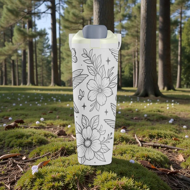 Pu&ograve; includere: Un bicchiere da viaggio bianco con un motivo floreale e alato nero. Il bicchiere ha un coperchio grigio e un bordo giallo chiaro. Lo sfondo &egrave; un'area erbosa verde con alberi.