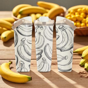 May include: Three white water bottles with a banana pattern, featuring a beige handle and lid. The bottles are on a wooden surface with bananas in the background. The bottles have the brand name "owala" printed on them.
