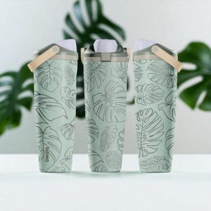 May include: Three water bottles with a pale green background and a repeating pattern of black monstera leaves. Each bottle has a white lid, a beige handle, and the brand name "owala" printed on the side.