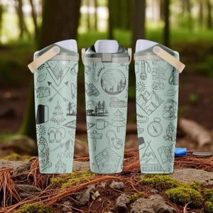 May include: Three light green insulated water bottles with black line art illustrations of camping and hiking themes. Each bottle has a white lid, a tan handle, and the word "ADVENTURE" on one. The bottles are on a natural forest floor.