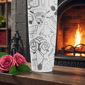 May include: A white tumbler featuring a repeating pattern of whimsical food and character illustrations in black. The tumbler is set on a wooden surface with two pink roses and green leaves. A fireplace with a burning fire is in the background.