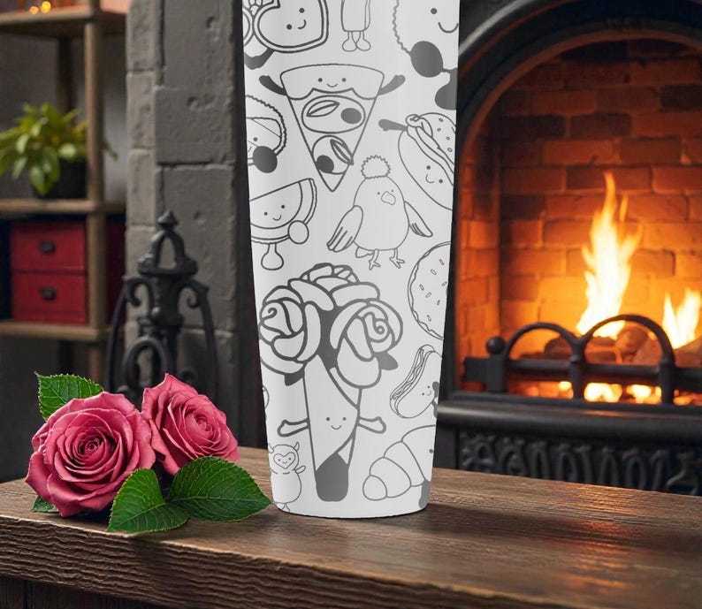May include: A white tumbler featuring a repeating pattern of whimsical food and character illustrations in black. The tumbler is set on a wooden surface with two pink roses and green leaves. A fireplace with a burning fire is in the background.