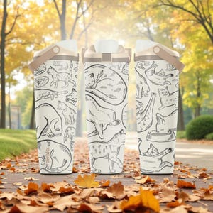 Peut inclure: Trois bouteilles d'eau isolées blanches avec un couvercle et une poignée beiges. Chaque bouteille présente un dessin au trait noir répété de chats dans diverses poses. Les bouteilles sont posées sur une surface en bois recouverte de feuilles mortes, avec un arrière-plan flou d'automne.