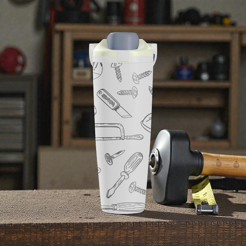 May include: A white water bottle with a gray lid and a pattern of hand-drawn tools, including screws, a saw, and a screwdriver. The bottle is cylindrical and sits on a wooden surface.