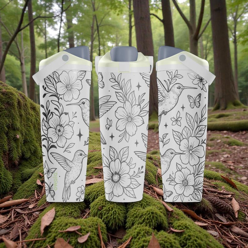 Pu&ograve; includere: Tre tazze da viaggio bianche con un design floreale e colibr&igrave;. Le tazze hanno un coperchio e un manico grigi. Il design presenta illustrazioni in bianco e nero di fiori, colibr&igrave;, farfalle e stelle. Le tazze sono ambientate su uno sfondo naturale della foresta.