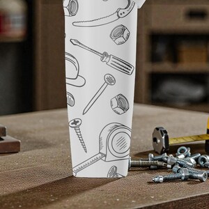 May include: A white tumbler featuring a repeating pattern of hand-drawn tools, including a saw, screwdriver, and tape measure. The tumbler is set on a wooden surface with additional tools and hardware in the background.